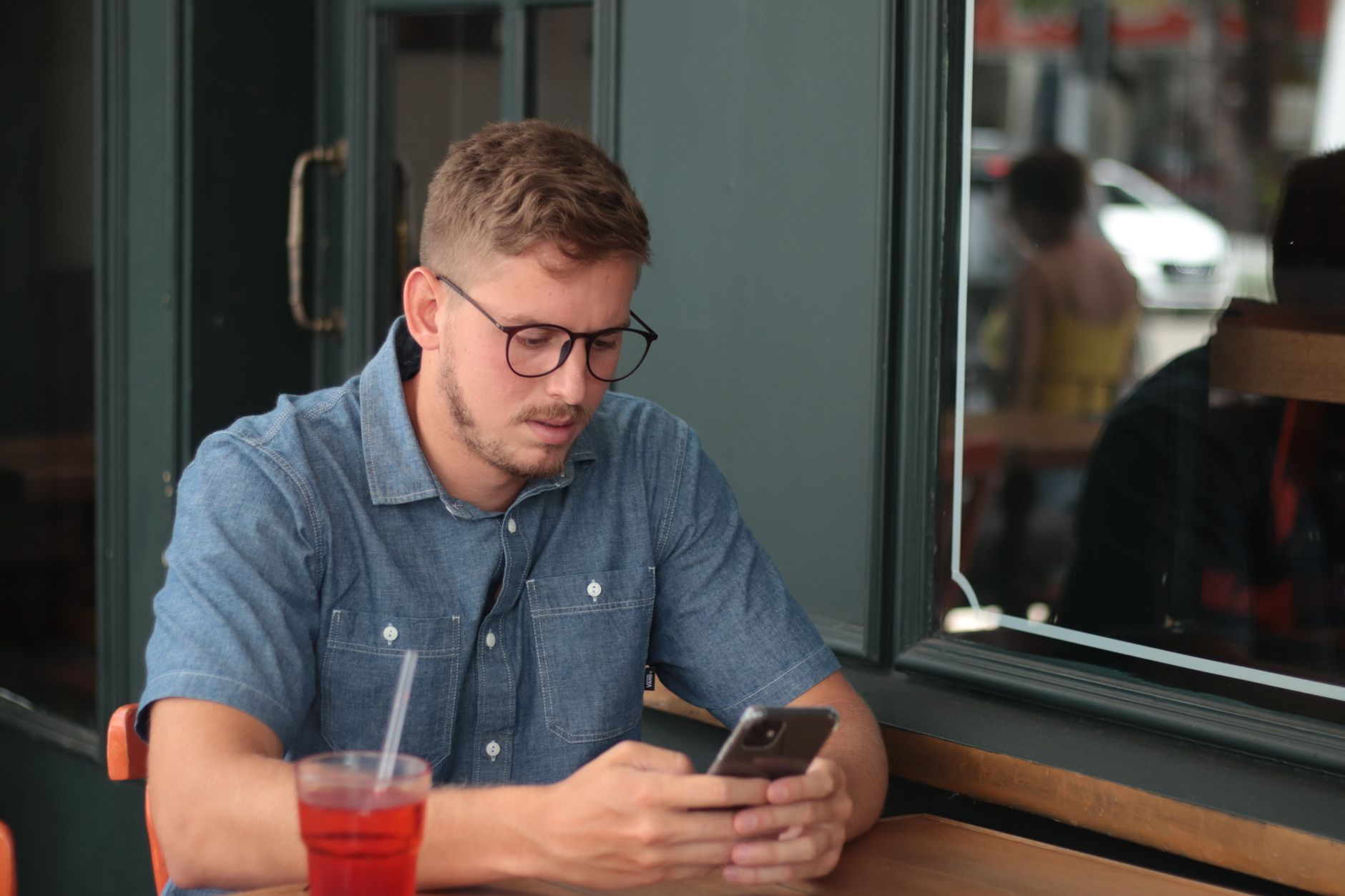 Un homme à lunettes consulte son smartphone à la terrasse d’un café, un verre posé devant lui