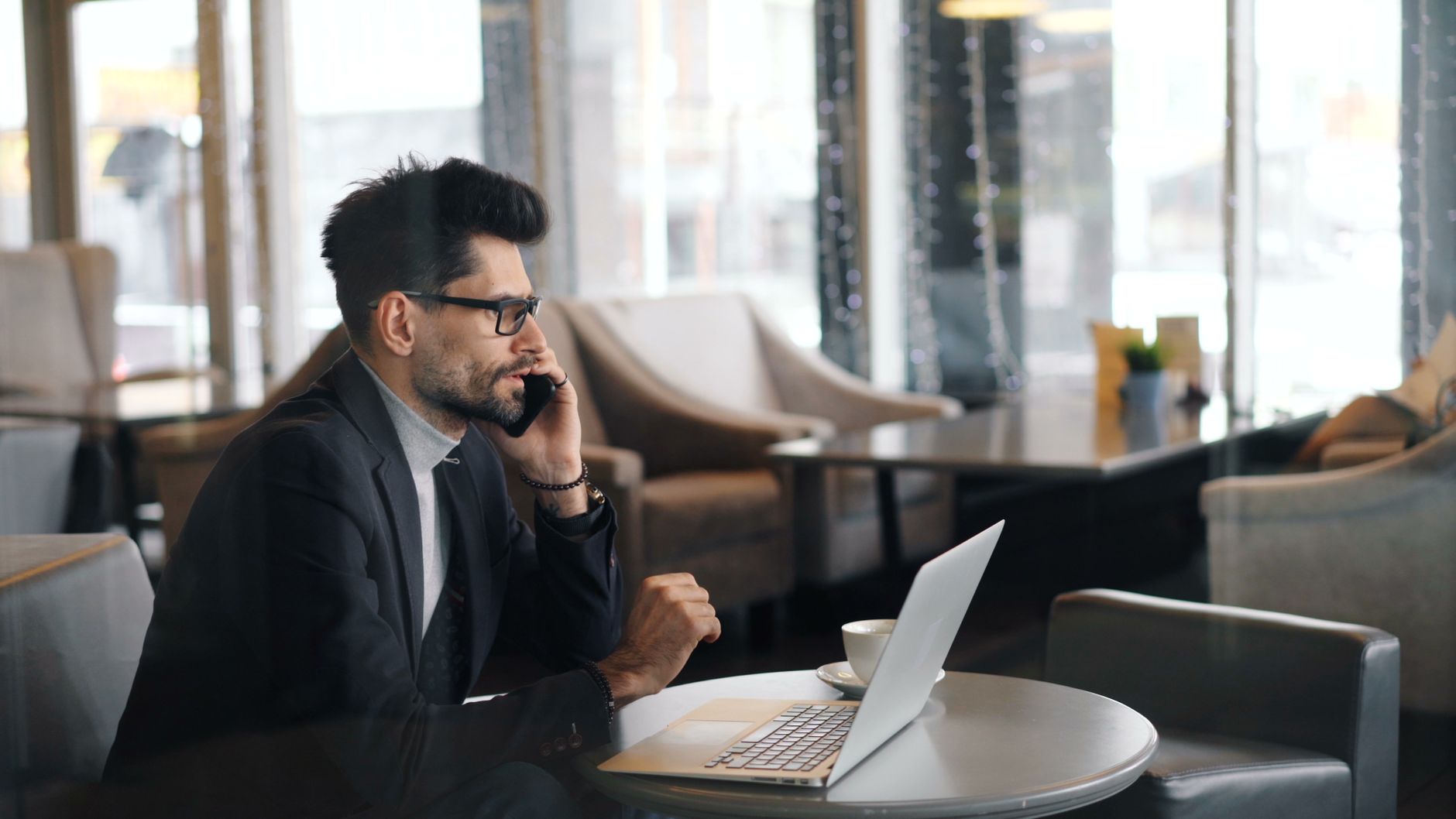 Un homme en veste sombre et lunettes téléphone dans un café, un ordinateur portable ouvert devant lui