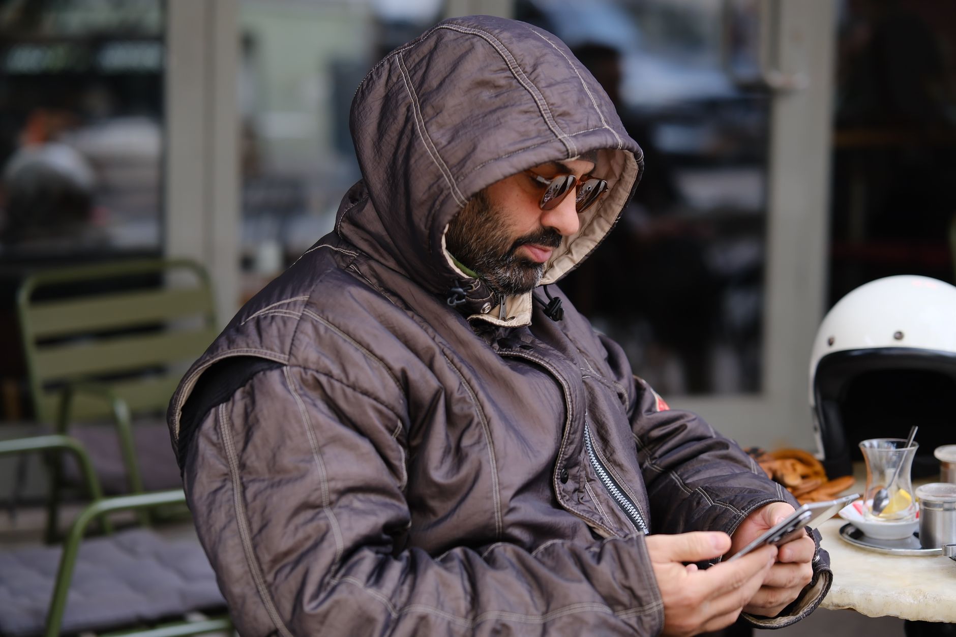 Un homme encapuchonné, lunettes de soleil sur le nez, regarde son smartphone à la terrasse d’un café